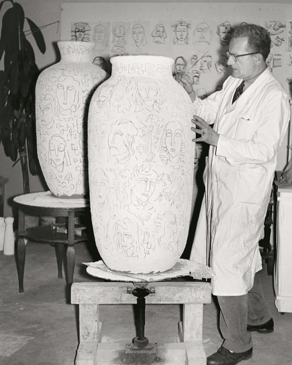 Ein Mann, gekleidet in einen weissen Arbeitskittel, bearbeitet eine hüfthohe Vase. Die Motive zeigen Gesichter. 