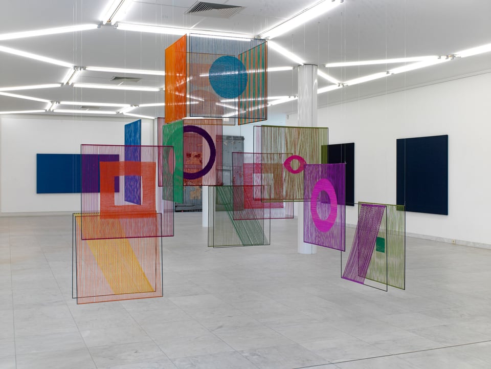 Ausstellungsansicht: In einem Raum hängen halbtransparente, quadratische und würfelförmige Elemente aus bunten Fäden bestehend. 
