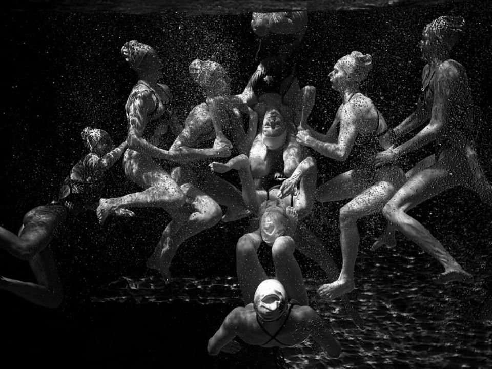 Ein knappes Dutzend junger Frauen in Badeanzügen hält sich unter Wasser fest und bildet eine Formation.