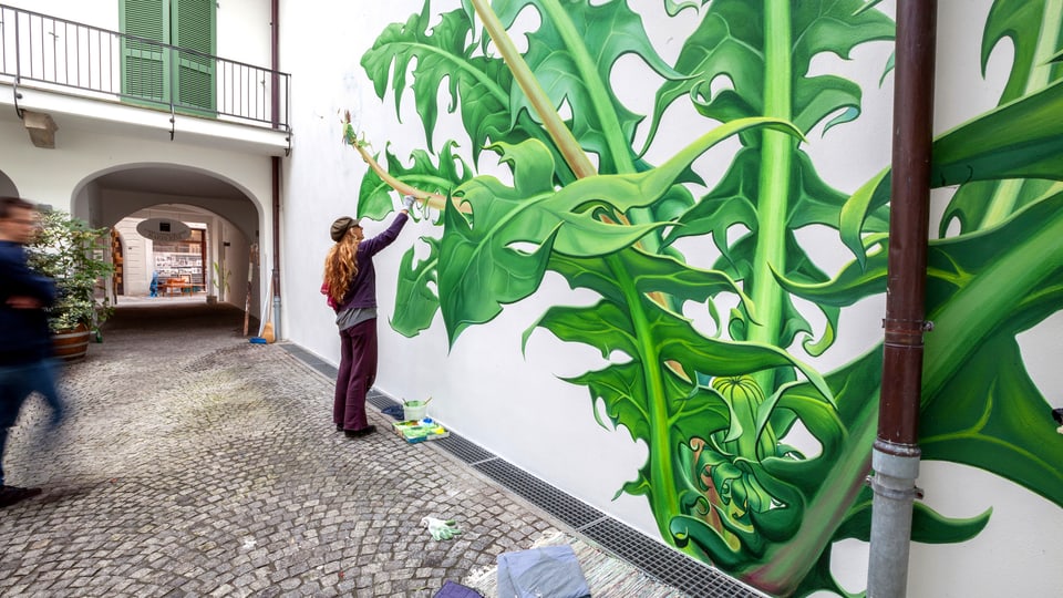 Eine Frau malt eine übergrossen Löwenzahn-Pflanze an eine Hausmauer.