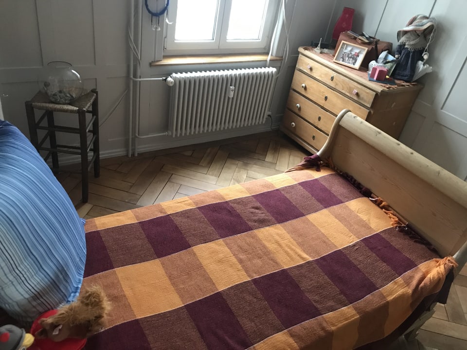 Ein Einzelbett aus Holz mit rot-oranger Decke steht in einem Gästezimmer. 