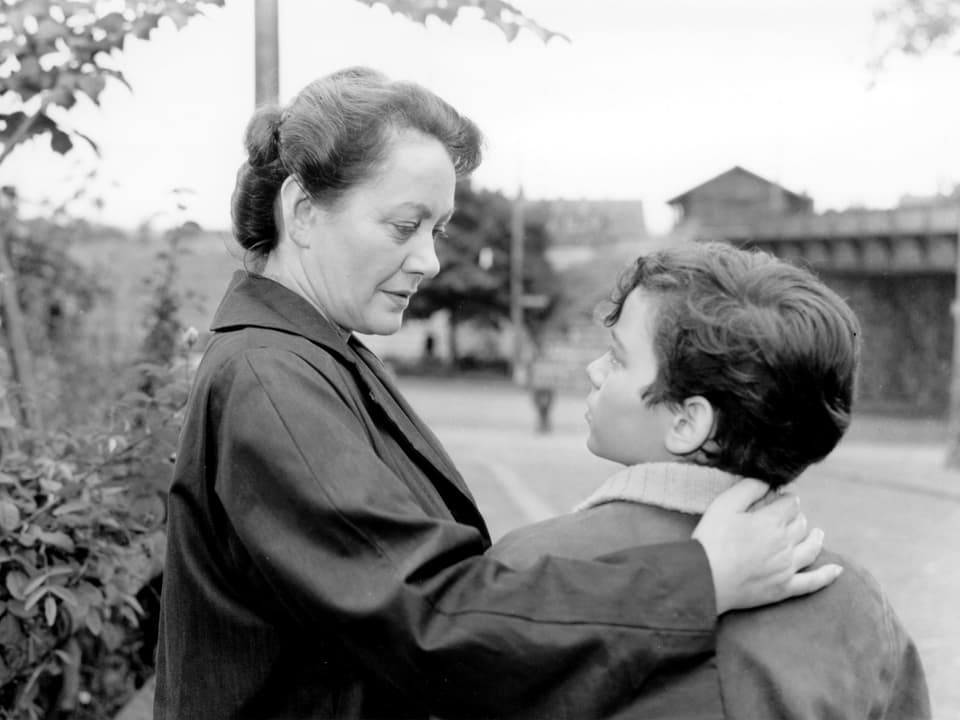 Eine Frau hält ihren Arm mütterlich um die Schultern eines Jungen, der zu ihr hinauf blickt.