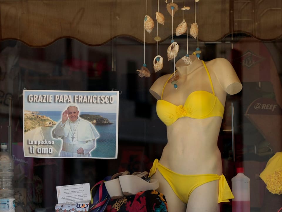 Zeitung mit Papstbild in einem Schaufenster neben einer Puppe mit Bikini.
