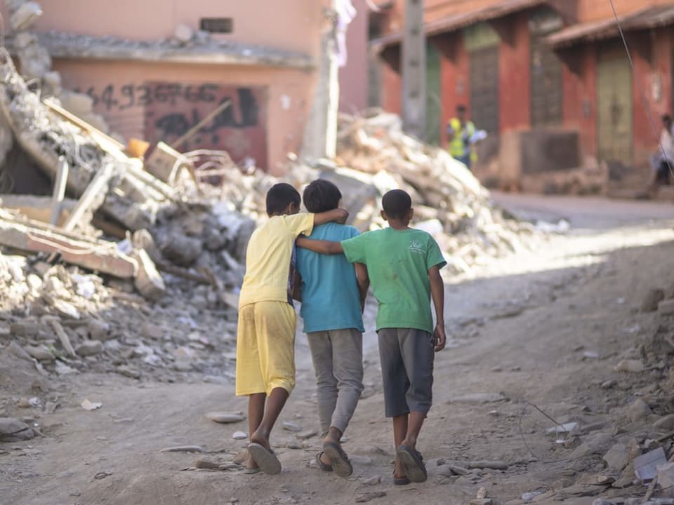 Kinder, fotografiert von hinten, halten sich gegenseitig fest, während dem sie durch die Trümmer ihrer Stadt laufen.