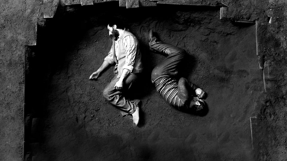 Zwei Männer liegen gekrümmt in einem Loch in der Erde.