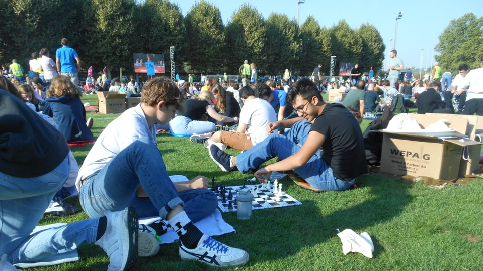 Man sieht zwei Jugendliche, die Schach spielen. Sie sitzen auf dem Fussballfeld auf dem Bode. 