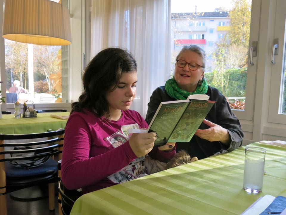 Eine Rentnerin liest mit einer Schülerin an einem Tisch ein Buch.