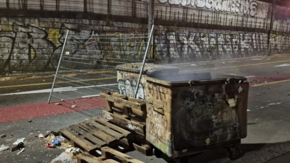 Müllcontainer und Paletten auf einer Strasse mit Graffiti-Wand im Hintergrund.