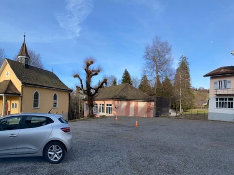 Links eine kleine Kapelle, in der Mitte ein rosa Haus (eine Turnhalle) und rechts der Ansatz eines weiss-rosa Hauses.