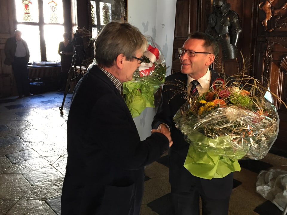 Zwei Politiker schütteln die Hand mit Blumensträussen in der Hand