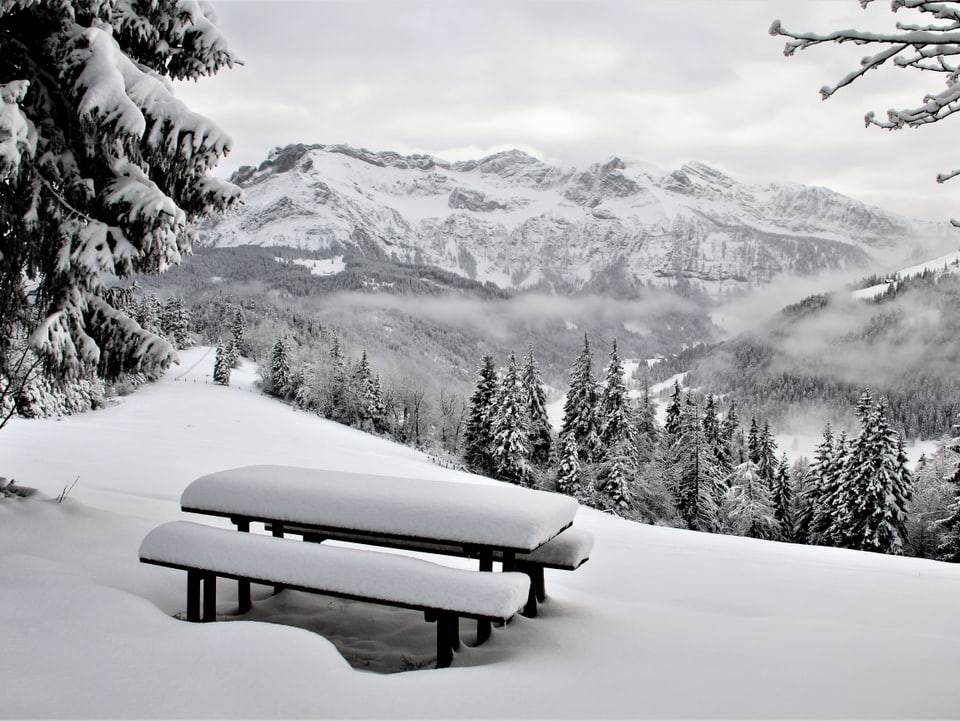 Im Vordergrund verschneiter Picknickplatz, dahinter Wälder und Hügel im Schnee.