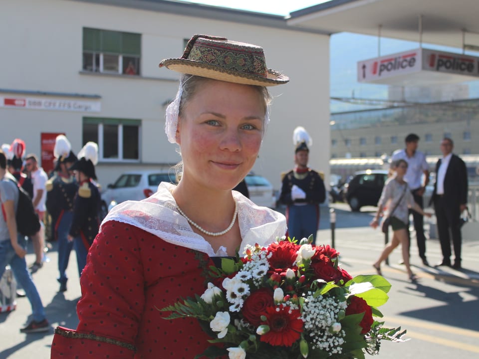 Frau mit Hut, rot-weissem Kleid und Blumenstrauss vor dem Sittener Bahnhof.