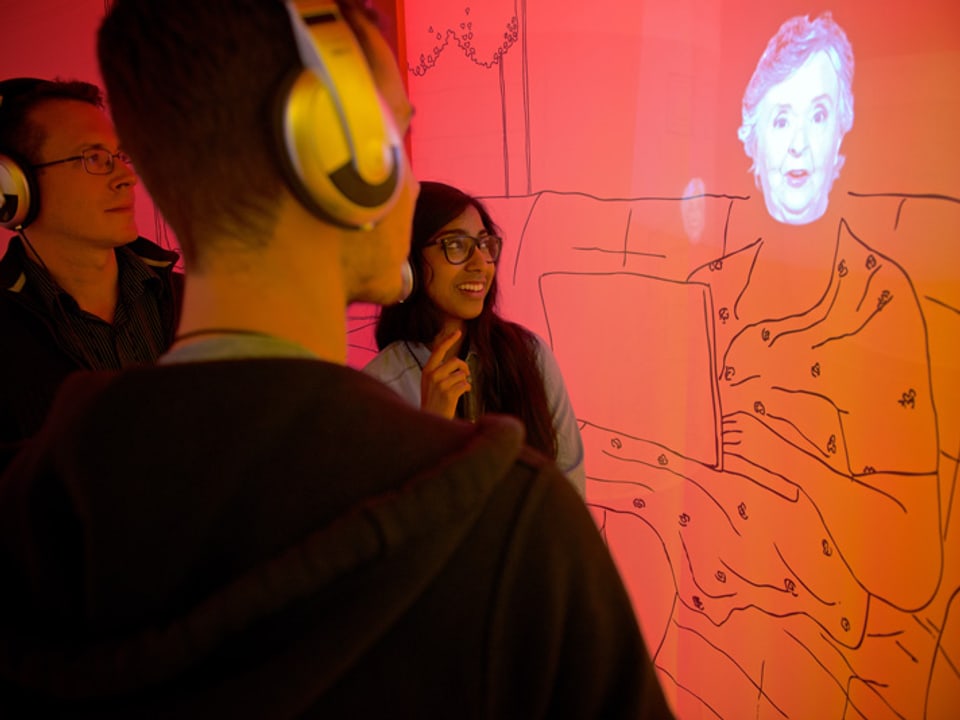 Museumsbesucher lauschen dem Audiokommentar einer Seniorin an einem Ausstellungsobjekt.