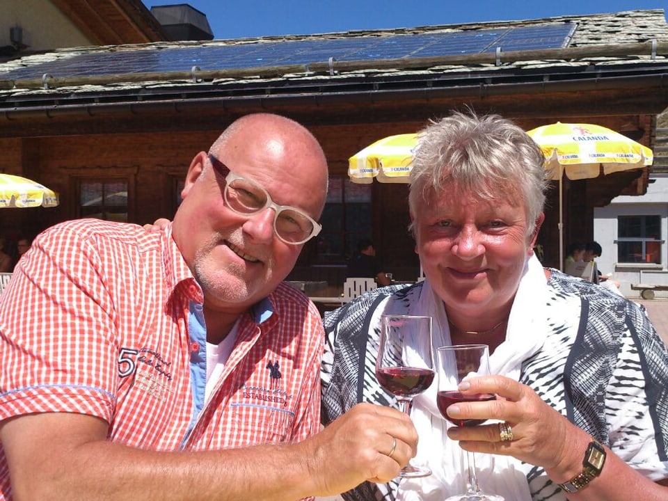 Ehepaar in der Sonne vor einer Bergwirtschaft, beide lachen und stossen mit einem Glas Rotwein an.