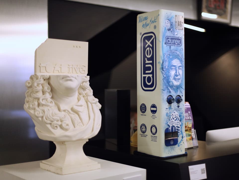 Ein besprühter Condom-Automat steht neben einer Skulptur im Museum.