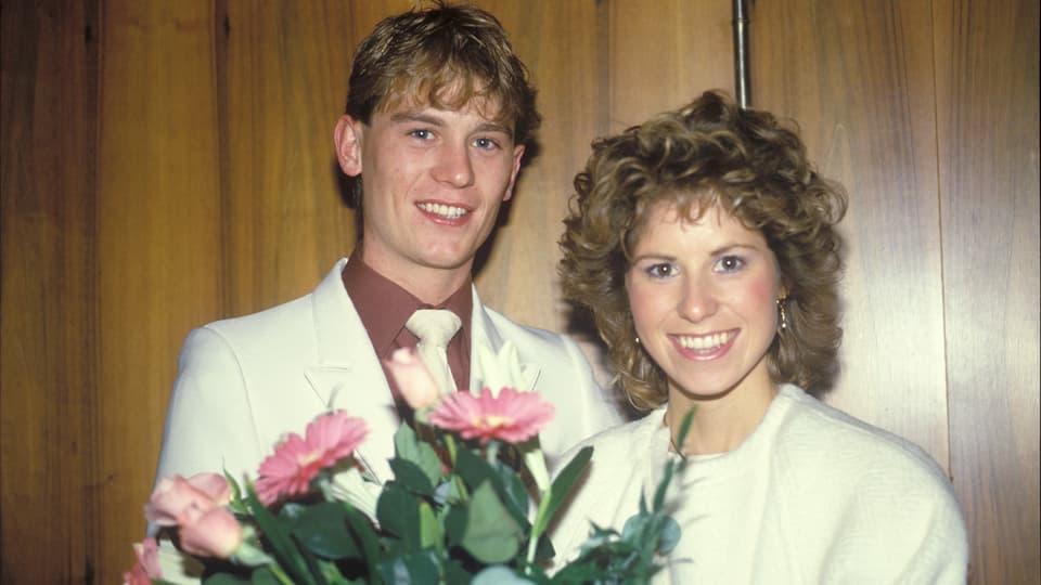 ein junges Ehepaar lächelt in die Kamera und hält einen Blumenstrauss