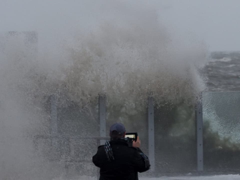Hohe Welle trifft auf Küste, davor steht ein Mann, der fotografiert.
