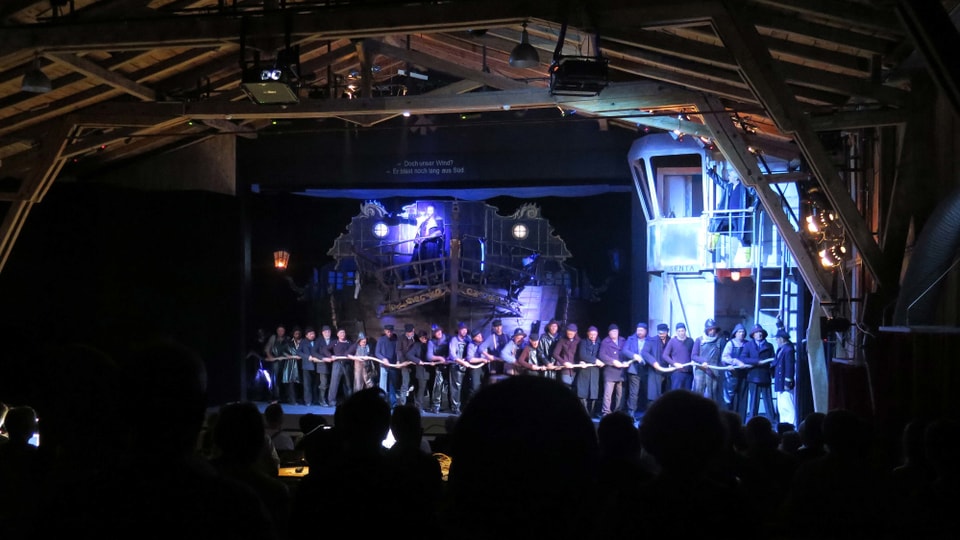 Chor aus Seemännern zieht an einem Seil, im Hintergrund das imposante Bühnenbild