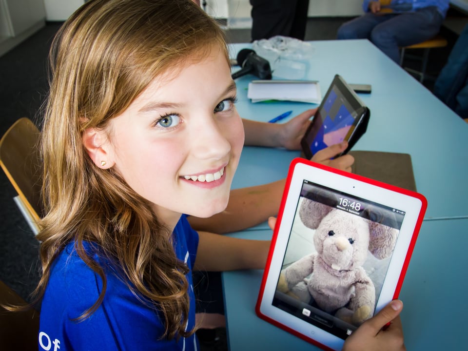 Ein zwölf jähriges Mädchen zeigt lachend ihren Tablet-Computer; darauf das Foto ihres Kuscheltieres.