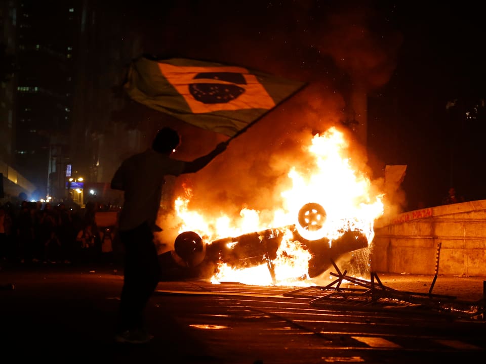 Nachtaufnahme: Im Hintergrund brennt ein Auto, davor schwenkt ein Demonstrant eine Brasilien-Flagge-