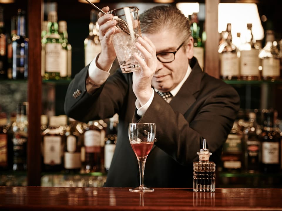 Ein Mann hält ein Glas mit Eiswürfeln über einem Cocktailglas mit rotem Inhalt. 