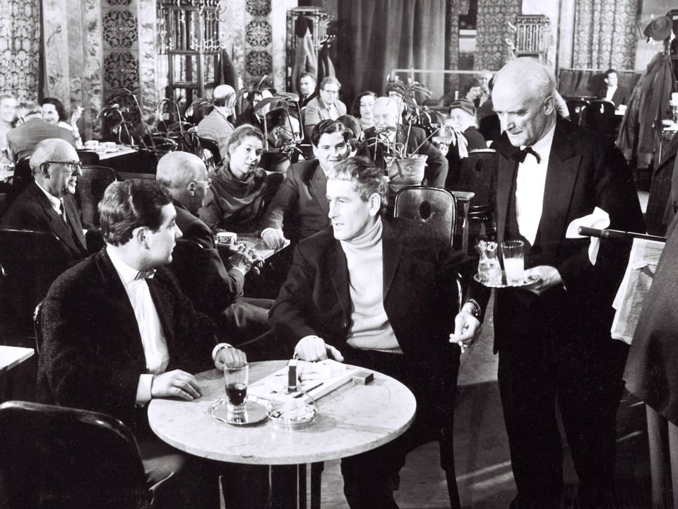 In einem vollen Café serviert der Kellner zwei Herren an einem runden Tisch im Vordergrund Getränke.