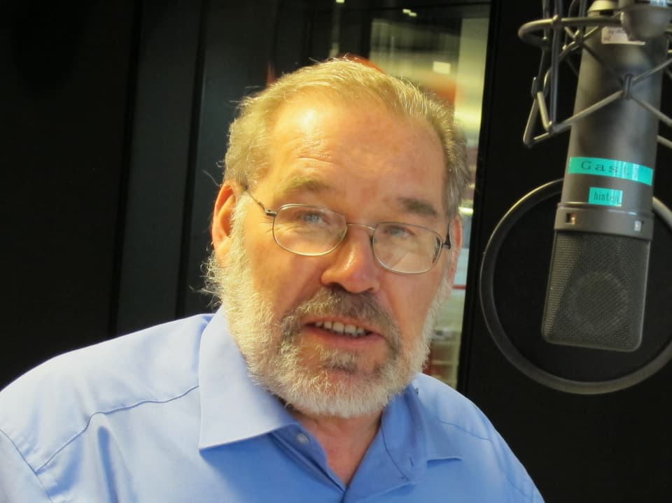Mann mit Brille und Bart und blauem Hemd neben einem Mikrofon.