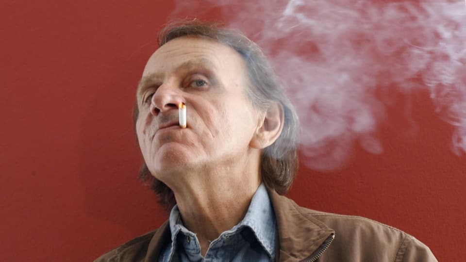 Ein Mann mittleren Alters mit schütterem Seitenscheitel schaut grimmig. Er hat eine Zigarette im Mundwinkel.