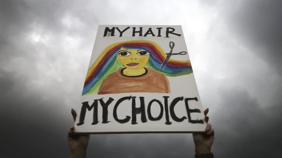 Ein Transparent wird gezeigt. Darauf: Eine Schere, die das regenbogenfarbige Haar abschneidet. My Hair, my choice.