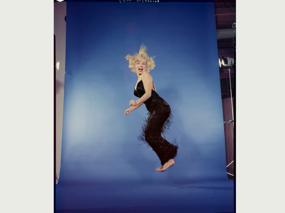Marylin Monroe springt vor blauer Leinwand in schwarzem Kleid. An den Seiten sind Studioleuchten sichtbar.
