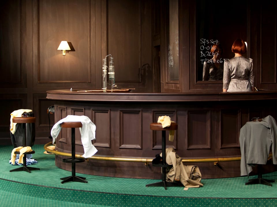 Bar in dunklem Holz, grüner Teppich, Bardame steht dahinter, auf den Barstühlen: Kleidungsstücke.