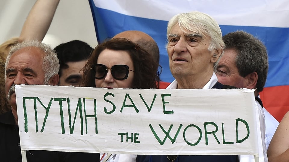 Russen vor einer grossen Landesflagge halten ein Schild hoch: «Putin rette die Welt» 