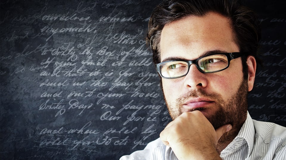 Bärtiger Mann mit Brille in Denkepose vor beschriebener Wandtafel