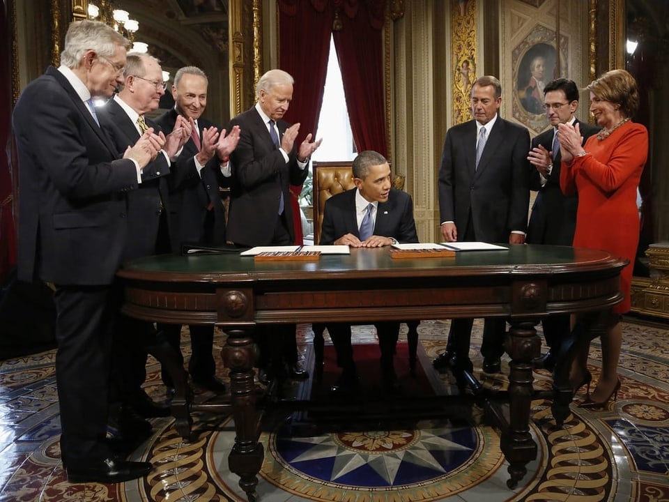 Barack Obama sitzt an einem Tisch. Um ihn herum applaudieren verschiedene Abgeordnete.