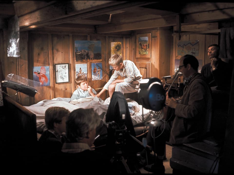 Zwei Kummerbuben Sitzen im Pijama auf dem Bett. Einer von ihnen auf der Lehne. Im Vordergrund Filmscheinwerfer und die Crew.