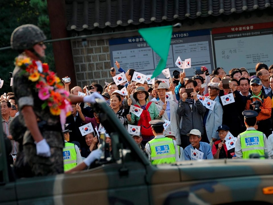 Ein Soldat in einem Militärfahrzeug trägt eine bunte Blütenkette, hinter ihm tobt die Menge und schwenkt voller Stolz die Fahne Südkoreas. 
