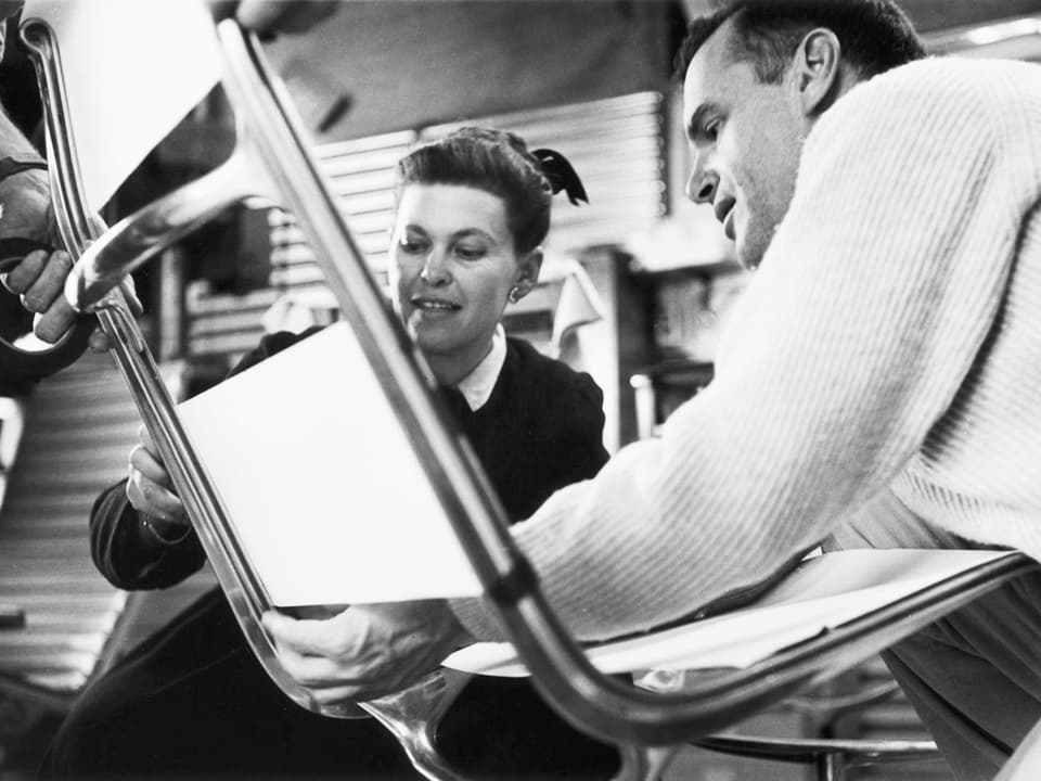 Im Vordergrund ist ein Stuhl aus Aluminium mit weisser Lehne und Sitzfläche zu sehen, dahinter und daneben Ray und Charles Eames, die den Stuhl überprüfen.