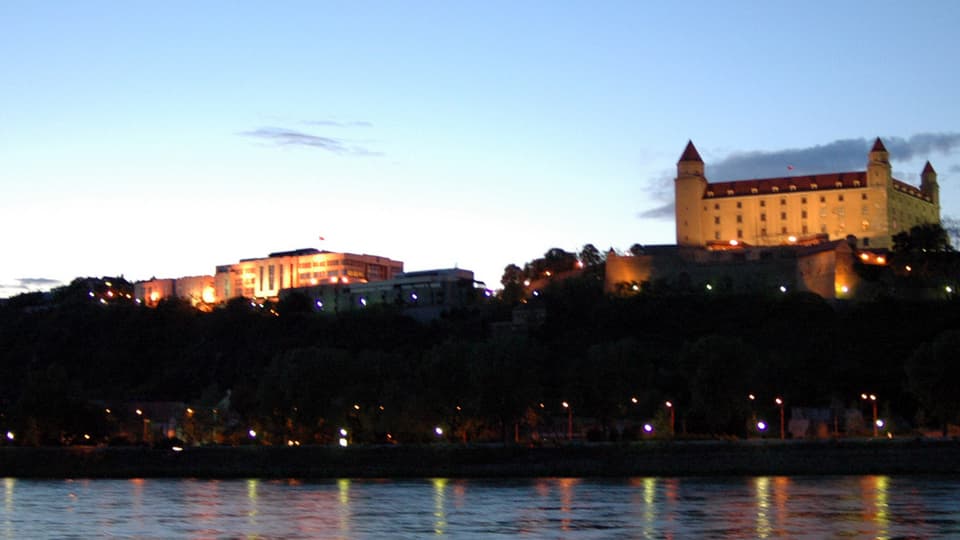 Die beleuchtete Burg in Bratislava in der Dämmerung, im Vorderund ein Fluss.
