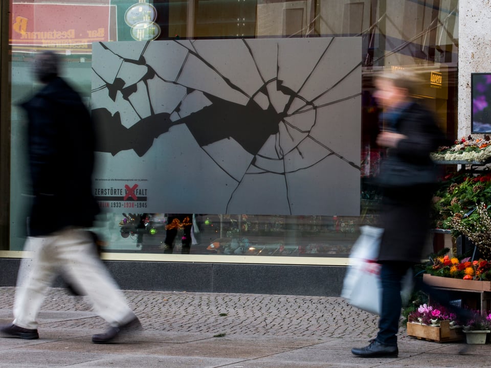 Leute laufen in Berlin an einem Geschäft vorbei, welches einen Aufkleber an der Scheibe hat, der zerbrochenens Glas symbolisiert.