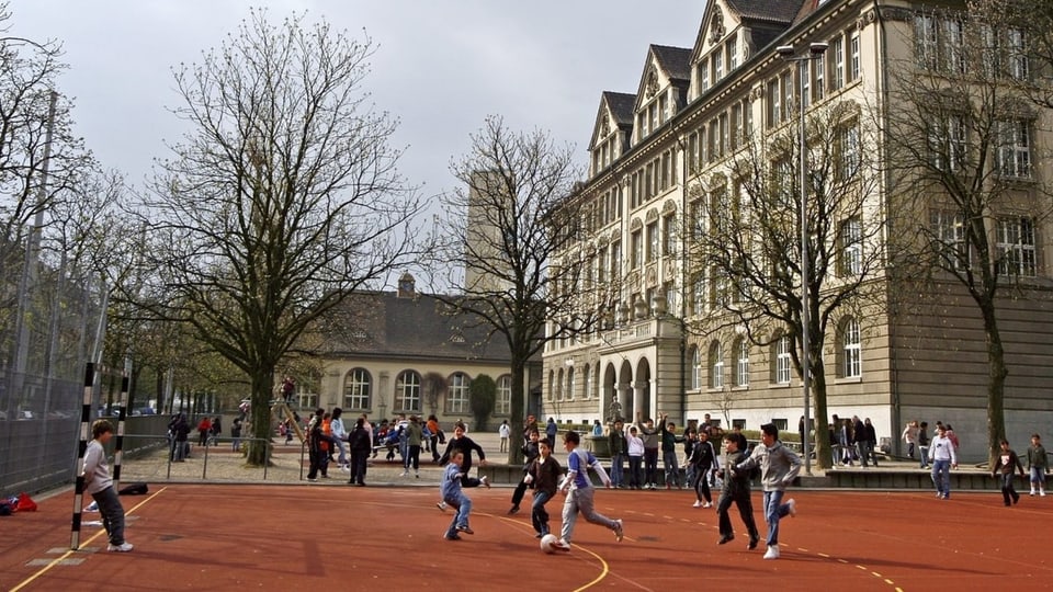 Kinder spielen auf einem Roten Platz vor einer Schule Fussball.
