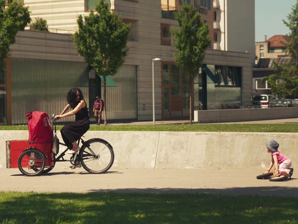 Eine Frau fährt mit dem Fahrrad, ein Kleinkind möchte ihr mit einem kleinen Fahrrad folgen