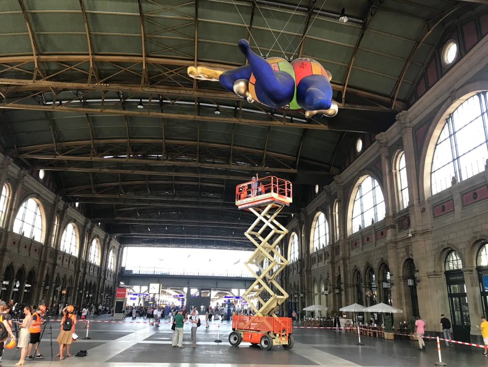 Ein Hebekran fährt langsam zu einem Kunstobjekt hoch, dass unter der Decke des Zürcher Hauptbahnhofs hängt.