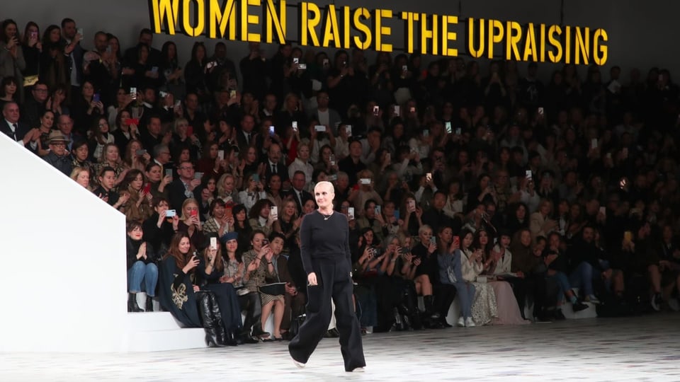 Frau an einer Modeschau. über ihr in grossen Buchstaben der Slogan «Women raise the upraising»