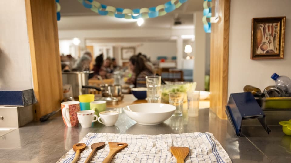 Eine Ablage mit Kochlöffeln, Geschirr und Gläsern. Im Hintergrund mehrere Personen (verschwommen) an einem Tisch.