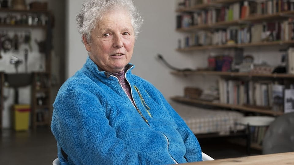 ältere Frau mit grauen kurzen Haaren, wachem Blick, blauem Pulli. Sie sitzt am Tisch und schaut in die Kamera.
