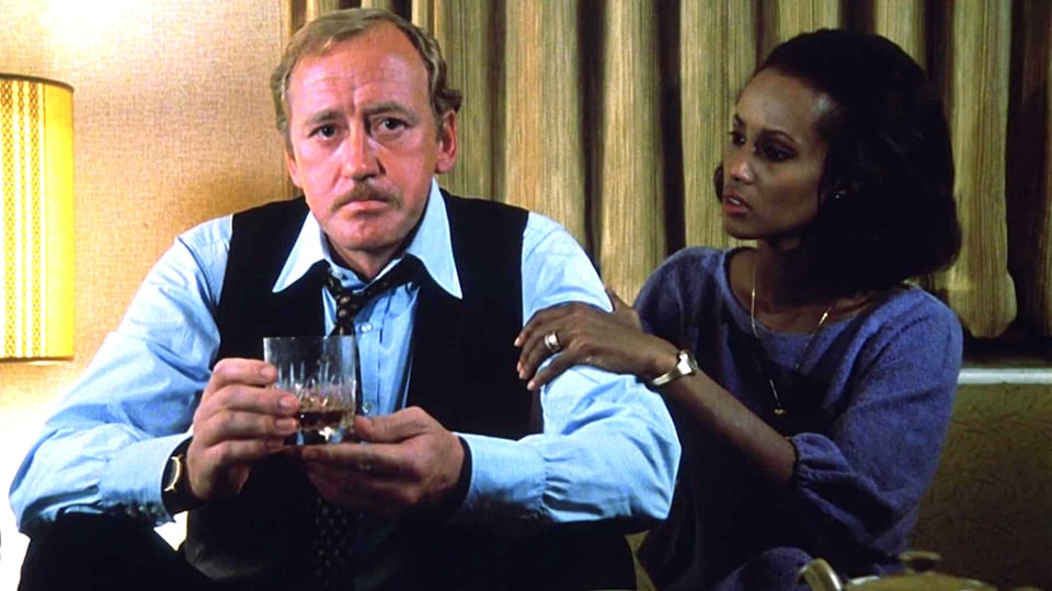 Ein Mann in Hemd, Krawatte und einem Glas Whisky in der Hand sitzt neben einer jungen Frau, die ihre Hand auf seinen Arm legt.