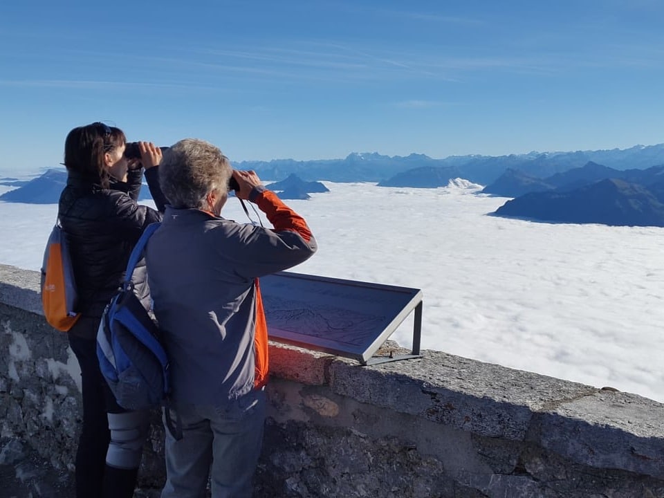 Zwei Frauen auf einem Berg mit Feldstecher bei Sonnenschein. Unten im Tal liegt Hochnebel. 