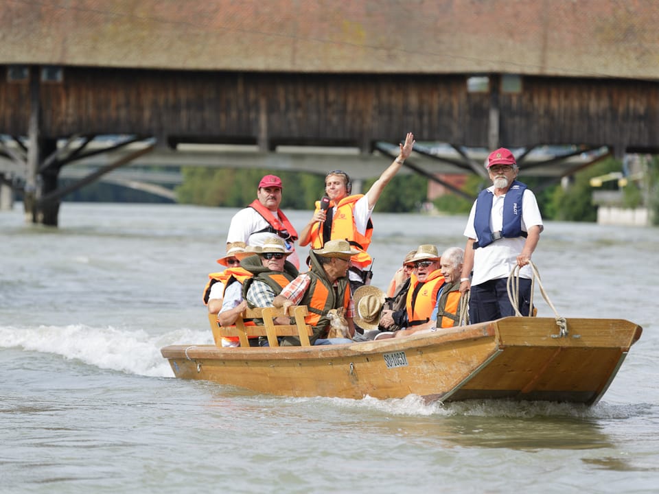 Reto Scherrer mit Mitwanderern im Boot, alle tragen Schwimmwesten. Im Hintergrund ist eine alte Holzbrücke zu sehen.