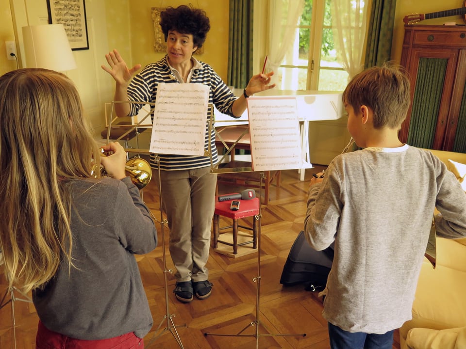 Eine Frau dirigiert, zwei Kinder spielen Trompete und Waldhorn.