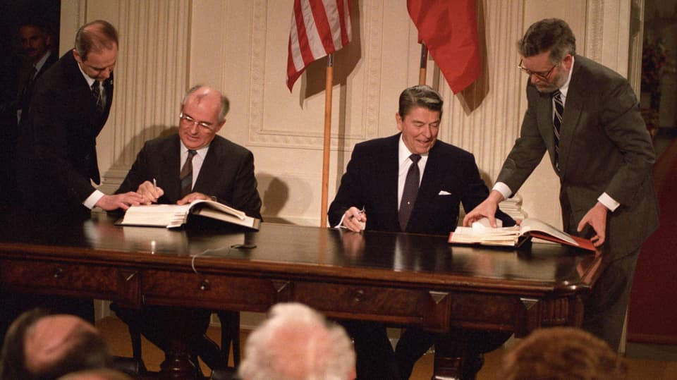 Zwei Männer sitzen vor der amerikanischen und sowjetischen Flagge und unterzeichnen Dokumente